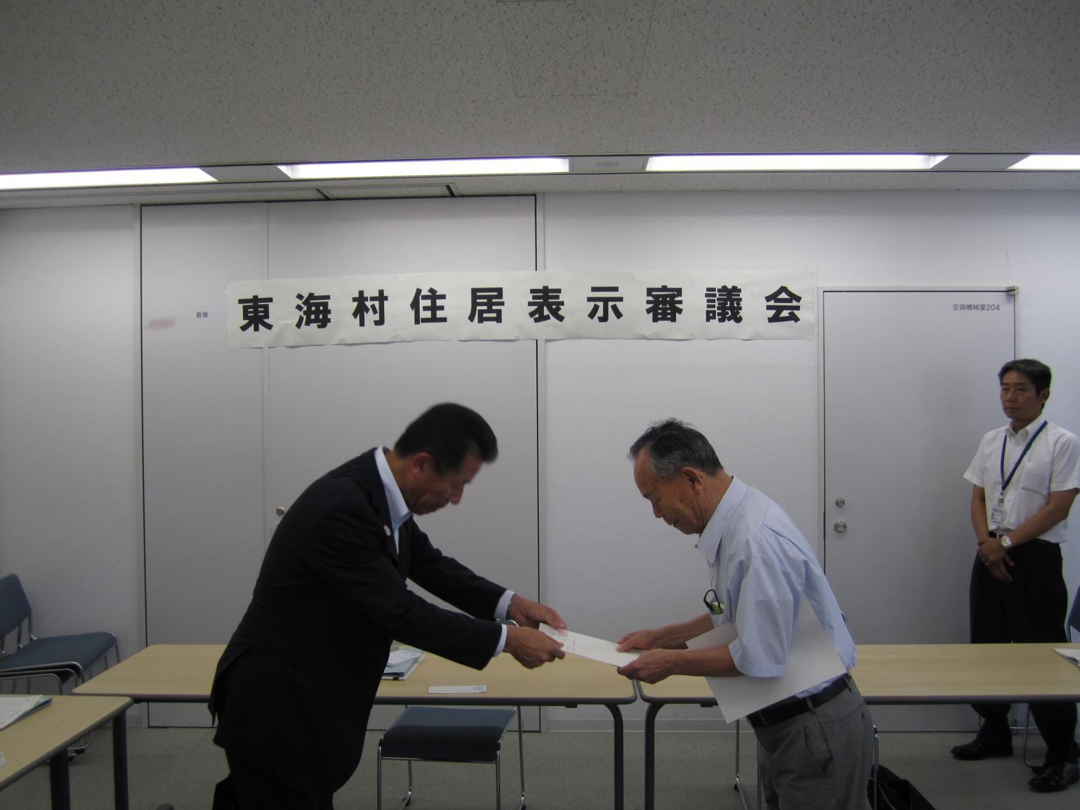 東海村住居表示審議会において、会議室にロの字型に並べられた机の内側で委嘱状を受ける寺門委員長の写真