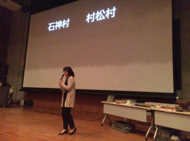 会場のスクリーンに、「石神村、村松村」と映し出され、その前でマイクを持って説明をする女性の写真