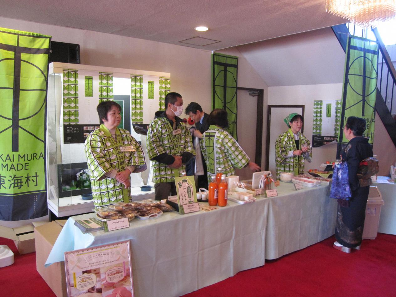 4人の黄緑色のはっぴを着た人がテーブルの上に置かれた東海村産の製品の前に立っている写真