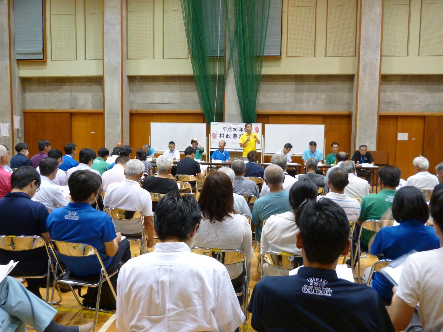 村松コミュニティセンターで行われた懇談会で、前方でマイクを持って話をしている男性と、着席し話を聞いている参加者たちの後方からの写真