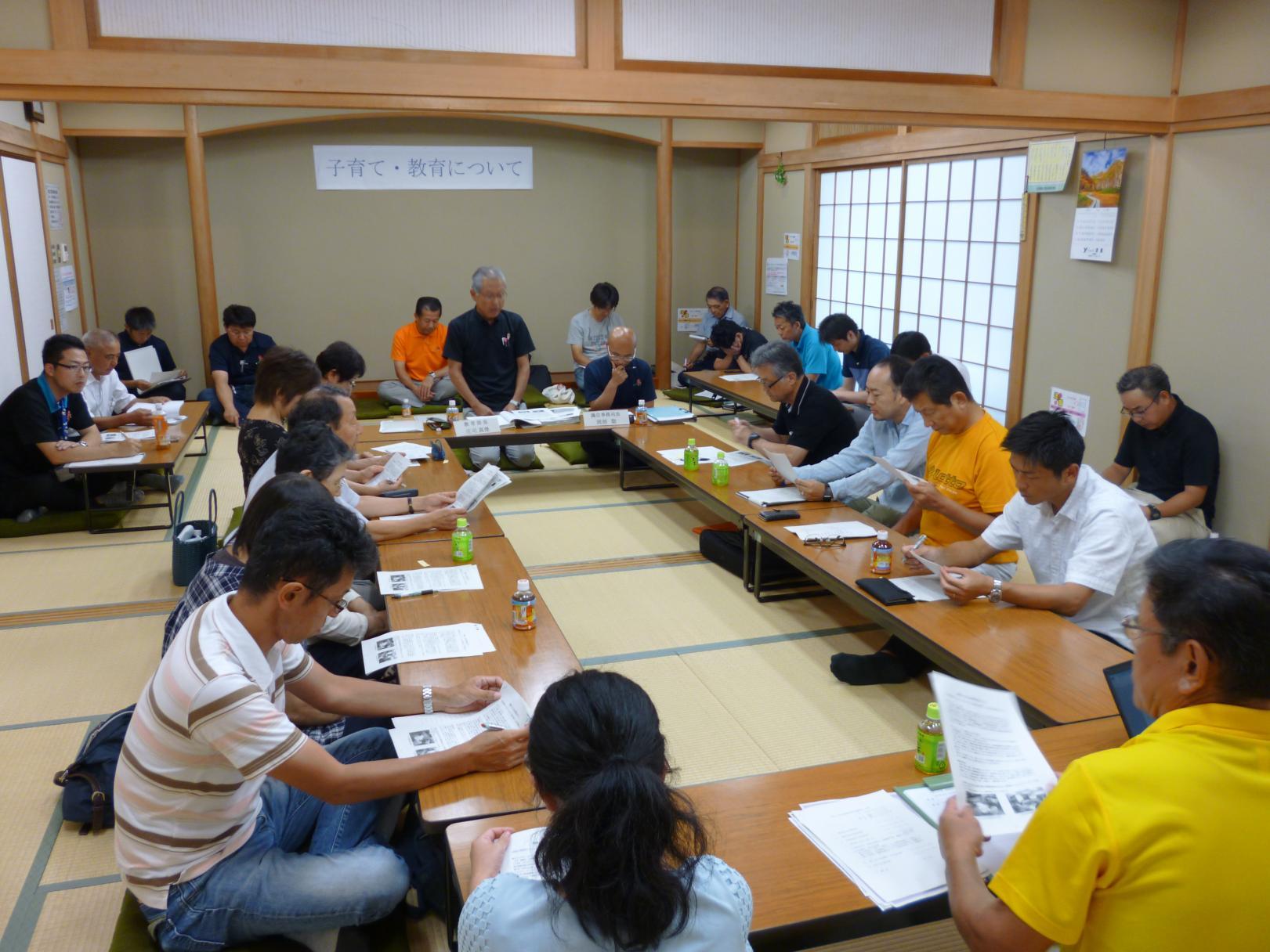 真崎コミュニティセンターで行われた懇談会(分科会)で、和室の部屋にロの字に配置された机の前に座って資料を見ている参加者たちの写真