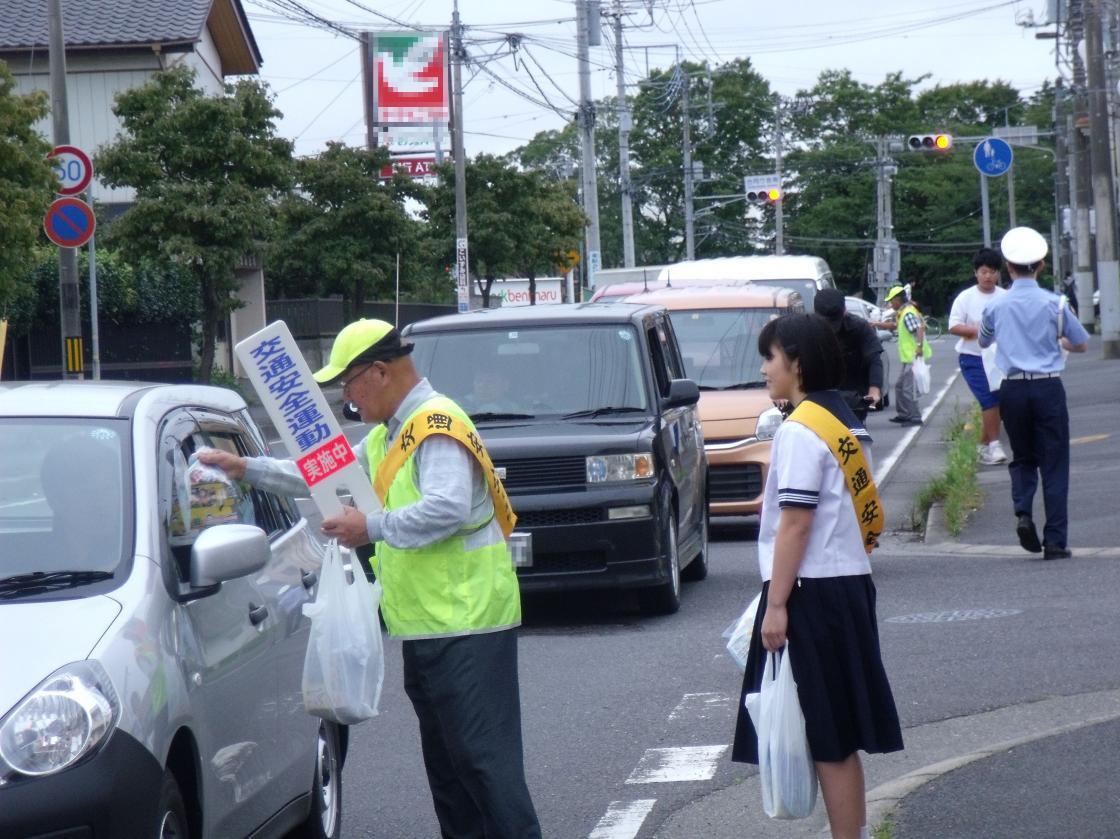 道路に停車中のくるまの助手席側から車の中へ、キャンペーンの広告などを手渡す男性と後ろに立つ女子学生の写真