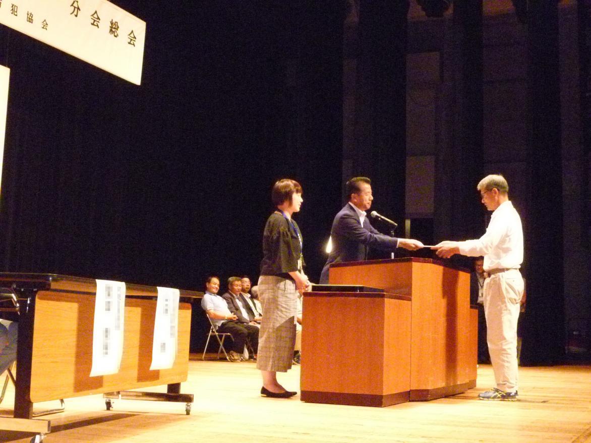 舞台の上で山田ひたちなか防犯協会副会長から委嘱状を受け取っている男性の写真