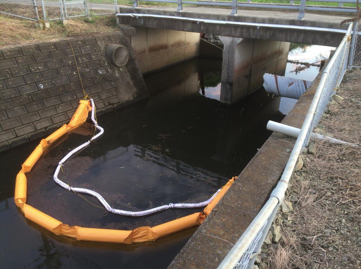 油の流れ込んだ水路の端から端に設置されたオイルフェンスの写真