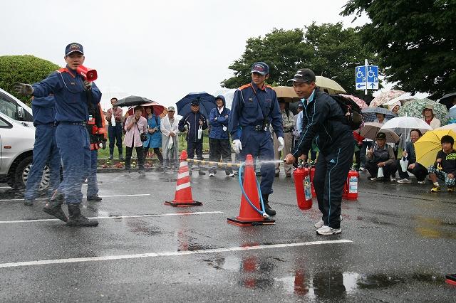 傘をさした参加者が見守る中、消防隊の方と消火器を実際に使った訓練をしている男性の写真