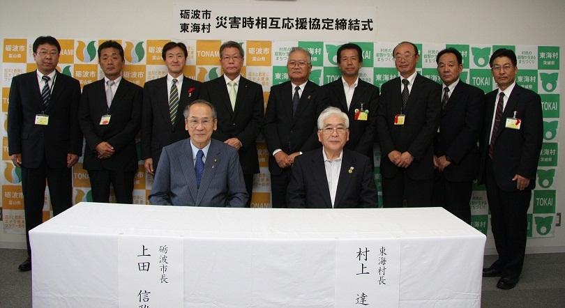 前列に上田砺波市長と村上東海村長が机の前で椅子に座り、後列に9人の男性が並んで立っている調印式後の集合写真