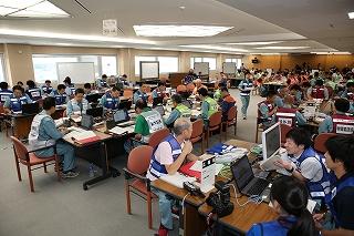 室内でグループごとにパソコンや資料の置かれた机で席に着き、体制や対応についての確認をしている訓練参加者たちの写真