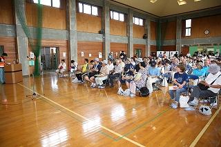 体育館に並べられた椅子に座っている参加者たちと前でマイクを持って説明している人の写真