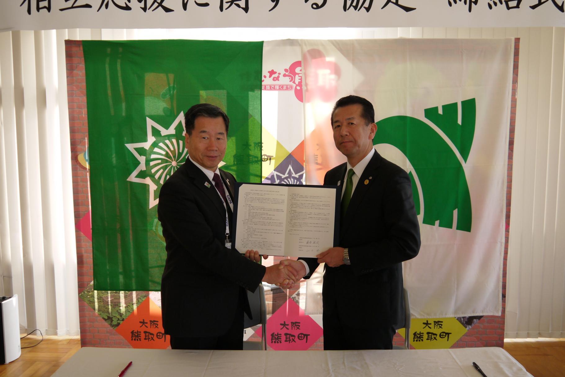 室内で協定書を2人で持ち、握手を交わす熊取町の藤原町長と山田村長の写真
